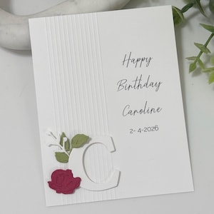 May include: A white birthday card with the text "Happy Birthday Caroline" and the date "2-4-2026". The card features a decorative "C" with a red rose and green leaves.