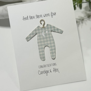 May include: A white card with the text "And then there were four" and a blue and white gingham baby onesie with a crescent moon and star design. The card also says "Congratulations Carolyn & Alex".