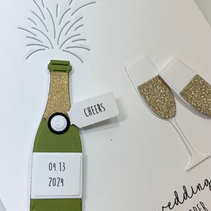 May include: A white card with a green and gold champagne bottle, two champagne flutes, and a cork popping out of the bottle. The card says "Cheers" and "04.13.2024" on the bottle. The text at the bottom of the card says "To your wedding, Joann & Cooper."