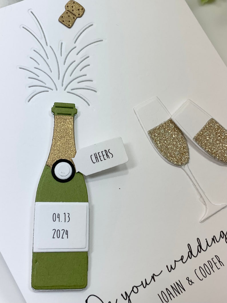 May include: A white card with a green and gold champagne bottle, two champagne flutes, and a cork popping out of the bottle. The card says "Cheers" and "04.13.2024" on the bottle. The text at the bottom of the card says "To your wedding, Joann & Cooper."