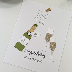 May include: A white card with a green champagne bottle, gold glitter champagne flutes, and the text "Congratulations on your engagement".