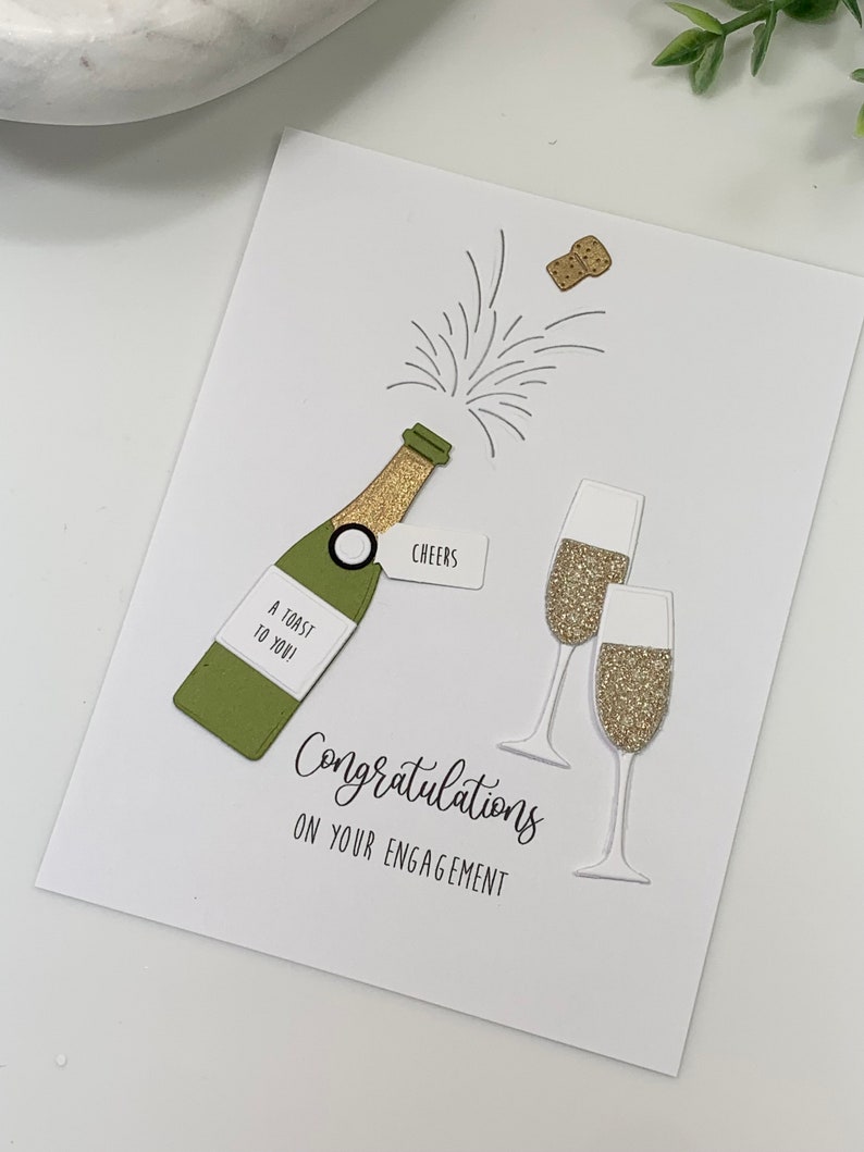 May include: A white card with a green champagne bottle, gold glitter champagne flutes, and the text "Congratulations on your engagement".