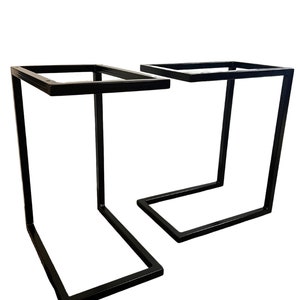 Puede incluir: Dos patas de mesa de metal negro con una parte superior cuadrada y una base rectangular. Las patas tienen forma de L invertida.