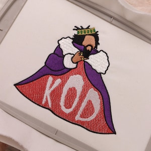 以下が含まれることがあります： 紫色のローブを着て金色の冠をかぶった王が、白い文字で「KOD」と書かれた赤い旗を持っている刺繍のデザインです。