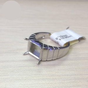 May include: A silver ring setting with a rectangular bezel and four prongs. The band features a series of vertical grooves. A white tag with text is attached to the ring.
