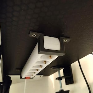 May include: A white power strip with multiple sockets is mounted under a dark surface. The power strip is held in place by a black bracket. The sockets have gold-coloured contacts. A red switch is visible on the power strip.