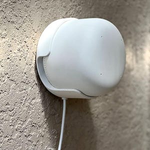 May include: A white, rounded wall-mounted device with a curved design. The device has a smooth surface and a small opening on the side. A white cord extends from the bottom. The device is mounted on a textured, light-colored wall.