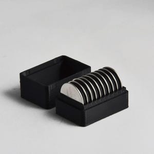 May include: A black rectangular box with a lid, containing several silver-colored, circular objects. The objects are stacked neatly inside the box. The box is open, revealing the contents. The background is plain white.