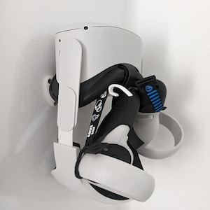 以下が含まれることがあります： 白いVRヘッドセットとコントローラーが壁掛けホルダーに取り付けられています。 ホルダーは白く、湾曲したデザインです。 黒いストラップとグリップがヘッドセットとコントローラーに取り付けられています。 「AMVR GRIP」の文字がストラップに表示されています。