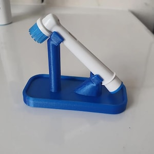 May include: A blue 3D-printed toothbrush holder with a white and blue electric toothbrush. The holder has a rectangular base and two supports to hold the toothbrush at an angle.