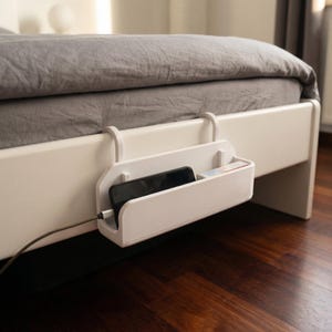 Puede incluir: Un organizador blanco para mesita de noche con un teléfono y un objeto pequeño dentro, colgado del marco de la cama. Un cable de carga está conectado al teléfono. La cama tiene una colcha gris.