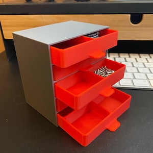 May include: A small, three-drawer storage unit with a silver top and gray sides. The drawers are bright red and partially open, revealing small metal objects. The unit is on a desk with a keyboard in the background.