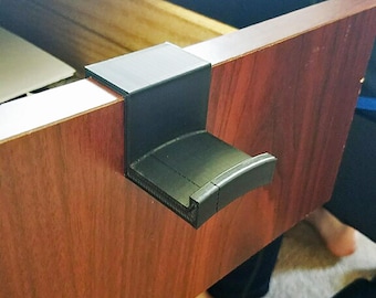 May include: A black headphone holder attached to a wooden desk. The holder is made of a matte black material and has a curved cradle to hold headphones. The desk is a dark wood color.