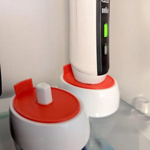 May include: White and red electric toothbrush and charging base. The toothbrush has a black panel with a green indicator light. The charging base is white with a red top. A separate white and red toothbrush head holder is also visible.