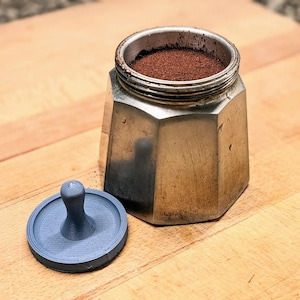 Peut inclure: Une cafetière italienne octogonale en argent remplie de café moulu, avec un tasseur bleu détaché sur une surface en bois. La cafetière est ouverte, révélant le café moulu brun foncé.