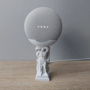 Può includere: Una statua bianca stampata in 3D di un uomo che tiene un altoparlante intelligente Google Home grigio su una base bianca.