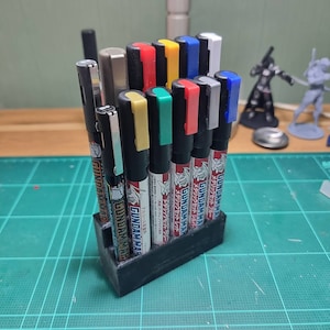 May include: A black plastic organiser holds a set of 12 Gundam Marker pens. The pens have black barrels with coloured caps and labels that read "Gundam Marker" in Japanese. The caps are red, yellow, green, blue, silver, and grey.