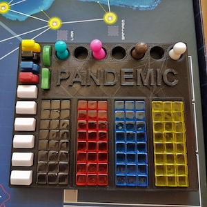 May include: A black game organiser for the board game Pandemic. The organiser has the word "PANDEMIC" printed on it. It holds coloured game pieces and cubes in various colours, including red, blue, yellow, black, and white. The game board is visible in the background.