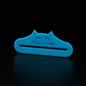 Puede incluir: Un exprimidor de tubos de pasta de dientes con forma de gato azul claro. El exprimidor tiene una ranura rectangular para el tubo y un diseño de cara de gato con orejas triangulares y ojos ovalados. El fondo es negro.