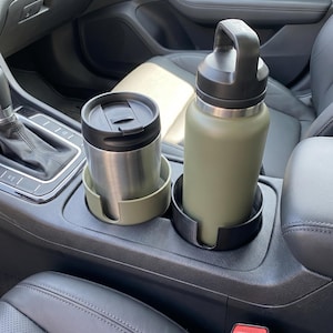 Può includere: Una tazza da viaggio in acciaio inossidabile e una borraccia verde oliva con manico nero sono nella console centrale di un'auto. Gli articoli sono in portabicchieri abbinati, uno verde chiaro e uno nero, progettati per le auto.