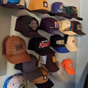 Könnte beinhalten: Eine Sammlung von Baseballkappen in verschiedenen Farben und Designs, darunter Schwarz, Orange, Braun und Blau. Einige Kappen zeigen Logos, Text und grafische Muster. Die Hüte werden an einer Wand ausgestellt.