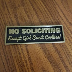 Puede incluir: Un letrero rectangular con las palabras "NO SOLICITING" en letras doradas sobre "Except Girl Scout Cookies!" en una fuente cursiva. El letrero tiene un fondo negro y un borde dorado.
