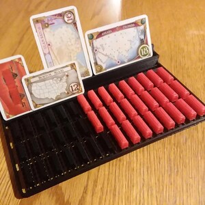 May include: A black game organiser with red game pieces and game cards. The cards have city names and numbers. The organiser has slots for the cards and pieces. The game pieces are red cylinders.