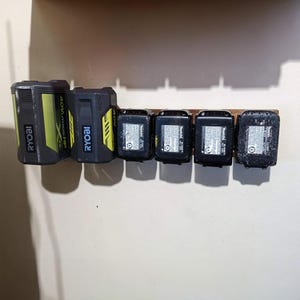May include: A wooden battery holder with six power tool batteries. Two Ryobi batteries are on the left, and four Makita batteries are on the right. The batteries are black and gray with yellow and white accents.