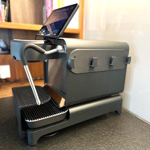 Puede incluir: Una máquina de espresso gris oscuro con una tableta integrada que muestra una interfaz digital. La máquina presenta una bandeja de goteo negra, una varilla de vapor plateada y un diseño elegante y moderno. La palabra "decent" está impresa en el lateral.