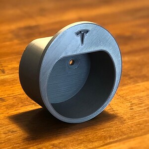 May include: A gray, 3D-printed Tesla emblem holder. The holder is cylindrical with a curved opening and a small hole. The Tesla logo is embossed on the top. The item is sitting on a wooden surface.