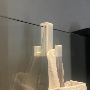 May include: A white plastic hook designed to hang over a glass panel. The hook has a rectangular shape with a curved top and two lower hooks. A white towel is hanging from the lower hook.