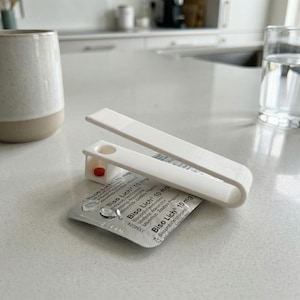 Peut inclure: Un distributeur de pilules blanc avec une pilule rouge à l'intérieur, posé sur une plaquette thermoformée de médicaments. La plaquette porte l'inscription "Biso Lich 10 mg". Une tasse en céramique et un verre d'eau sont en arrière-plan.