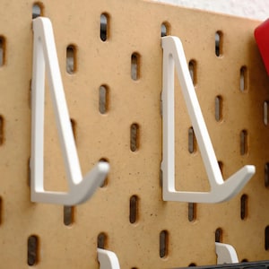 May include: Close-up of a pegboard with white metal shelf brackets. The brackets are angled and designed to hold items. The pegboard is a light brown color with evenly spaced holes.