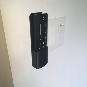 May include: A black remote control with white icons and buttons is mounted on a wall. The remote has a power button, volume controls, and other function buttons. A white light switch plate with two switches is next to the remote.