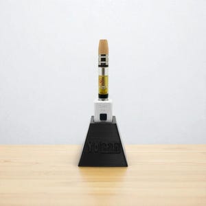 Peut inclure: Un support noir en forme de pyramide avec le texte "YoCan" maintient un stylo vape. Le stylo vape a un embout en bois, un corps argenté et une cartouche transparente remplie d'un liquide doré. Le support est sur une surface en bois.