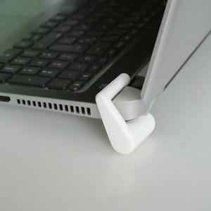 Könnte beinhalten: Ein weißer Laptop-Ständer, der einen Laptop stützt. Der Ständer besteht aus einem soliden, glatten Material und ist so konzipiert, dass er den Laptop für eine verbesserte Ergonomie anhebt. Die Tastatur des Laptops ist sichtbar.