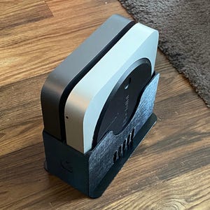 以下が含まれることがあります： 黒とシルバーのコンピューターデバイスが黒いスタンドに収められています。コンピューターは、シルバーの上面と黒いベースを備えた丸みを帯びた正方形の形状をしています。スタンドには一連の長方形の切り欠きがあります。デバイスは木製の表面にあります。