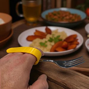 Può includere: Una forchetta d'argento con un'impugnatura gialla per le dita. L'impugnatura è progettata per aiutare a tenere le posate. Lo sfondo mostra un tavolo di legno con piatti di cibo e un bicchiere di liquido.