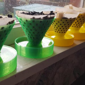 Peut inclure: Des jardinières coniques en plastique vert et jaune avec un motif quadrillé, contenant de la terre et des filtres en papier. Les jardinières sont posées sur un rebord de fenêtre, avec un arrière-plan flou.