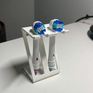 Puede incluir: Un soporte blanco para cepillos de dientes con dos cabezales de cepillo de dientes eléctricos. Los cabezales del cepillo de dientes tienen cerdas azules y verdes. El soporte está sobre una superficie blanca.