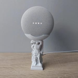 Può includere: Un altoparlante intelligente grigio è posto su una statua bianca di un uomo. L'altoparlante ha quattro punti luminosi ed è tenuto da un supporto curvo. La statua è dettagliata con figure classiche e poggia su una base rettangolare.