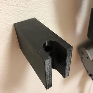 Puede incluir: Un soporte de pared rectangular negro con una abertura curva. El soporte está hecho de un material sólido y está montado en una pared de color claro. El diseño sugiere que es para sujetar un objeto.