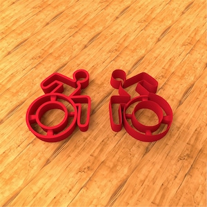 Pode incluir: Dois símbolos de cadeira de rodas vermelhos são exibidos em uma superfície de madeira. Os símbolos são estilizados, com uma roda circular e uma figura representando uma pessoa. O fundo é um grão de madeira marrom claro.