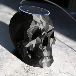 Könnte beinhalten: Ein schwarzer, geometrischer Lautsprecher in Schädelform mit einer kreisförmigen Oberseite. Der Lautsprecher hat ein facettiertes Design und ein blau leuchtendes Licht um die Oberseite. Ein schwarzes Kabel erstreckt sich von der Oberseite des Lautsprechers.