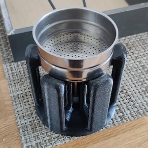Op de afbeelding: Een zilveren koffiefilter met een zwarte kunststof basis. Het filter heeft een geperforeerde metalen bodem en een gladde, afgeronde bovenkant.
