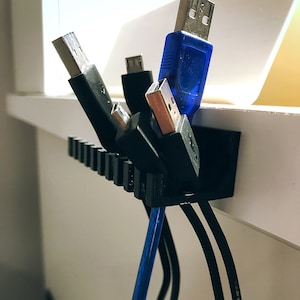 Könnte beinhalten: Ein schwarzer Kabelorganizer aus Kunststoff mit mehreren Schlitzen zum Halten von Kabeln und Drähten. Der Organizer ist an einer weißen Oberfläche montiert und hat mehrere Kabel eingesteckt, darunter ein blaues USB-Kabel.