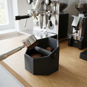 Op de afbeelding: Een zwarte, zeshoekige espresso afklopbak met een roestvrijstalen portafilter. De bak bevat koffiepucks en een grijze staaf. Een espressomachine en een molen staan op de achtergrond, alles op een houten aanrecht.