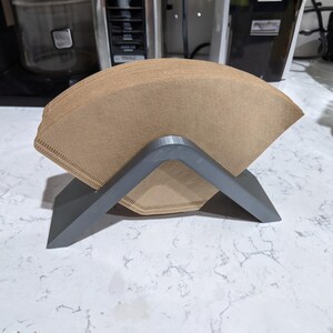 Può includere: Una pila di filtri per caffè in carta marrone appoggiata su un supporto grigio angolato. Il supporto ha un design semplice e moderno, con una forma triangolare. I filtri sono ben impilati, pronti per l'uso. Lo sfondo è un piano di lavoro della cucina.