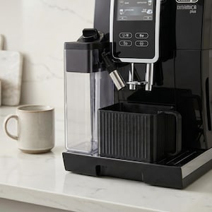 Könnte beinhalten: Eine schwarze De'Longhi Dinamica Plus Espressomaschine mit digitalem Display, Wassertank und schwarzer Tropfschale. Links von der Maschine steht eine beige Tasse auf einer weißen Arbeitsplatte.