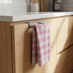 Può includere: Un portasciugamani grigio è montato su un mobile in legno, che tiene un canovaccio da cucina a quadri rosa e bianco. L'asciugamano è piegato e appeso. L'ambiente della cucina comprende un piano di lavoro bianco e mobili in legno chiaro.